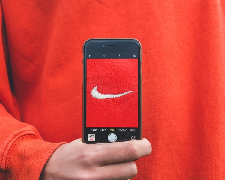 Nike-Logo in einem Handy von einer Hand vor einem roten Pullover gehalten